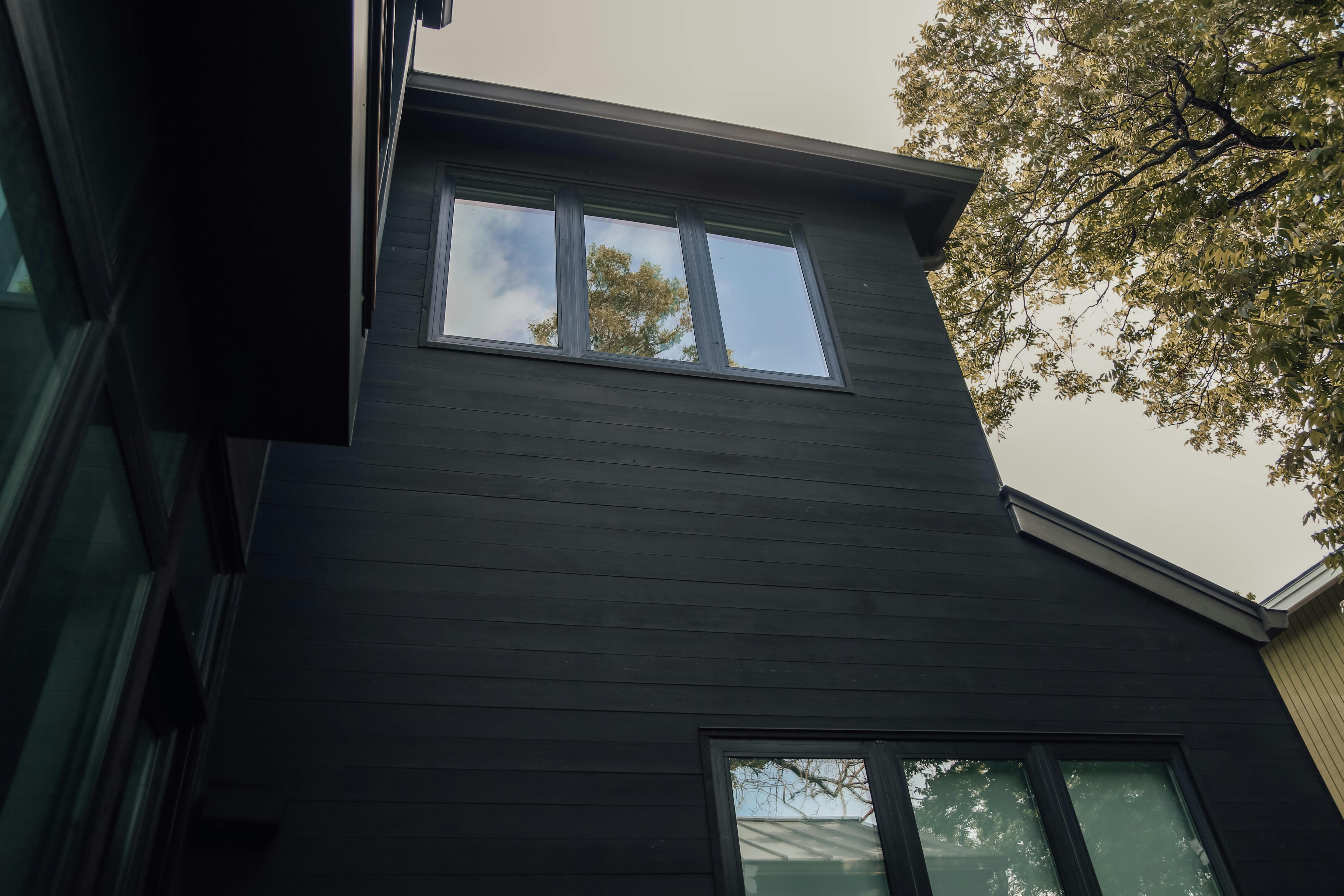 From below of gray sky and green foliage over tall black house with small windows and black wooden walls in daytime